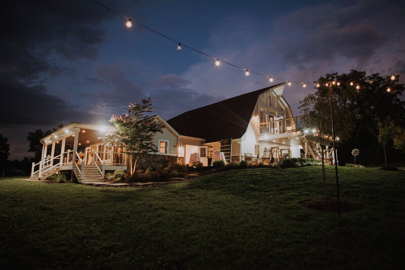 A warmly lit barn-style building at dusk, featured on https://halcyon-watson-1.showit.site/the-venue, boasts string lights above an outdoor area. With the deep blue sky and a large tree to the right, the scene exudes a cozy, inviting atmosphere against a grassy foreground.