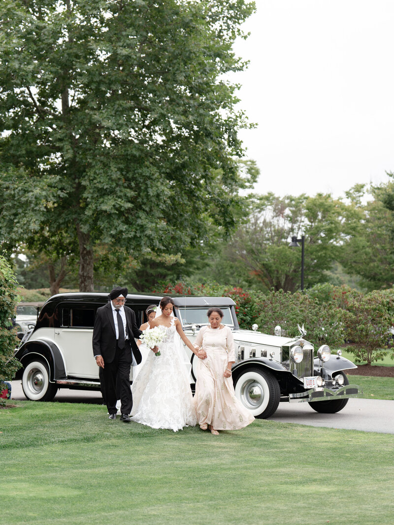 Bride walks with her parents towards ceremony with a Rolls Royce behind them