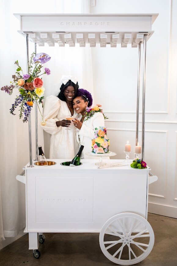 A couple in wedding attire cheers glasses of champagne while embracing behind a cart with a two tier floral wedding cake, bottles of champagne, and floral and candle decorations.