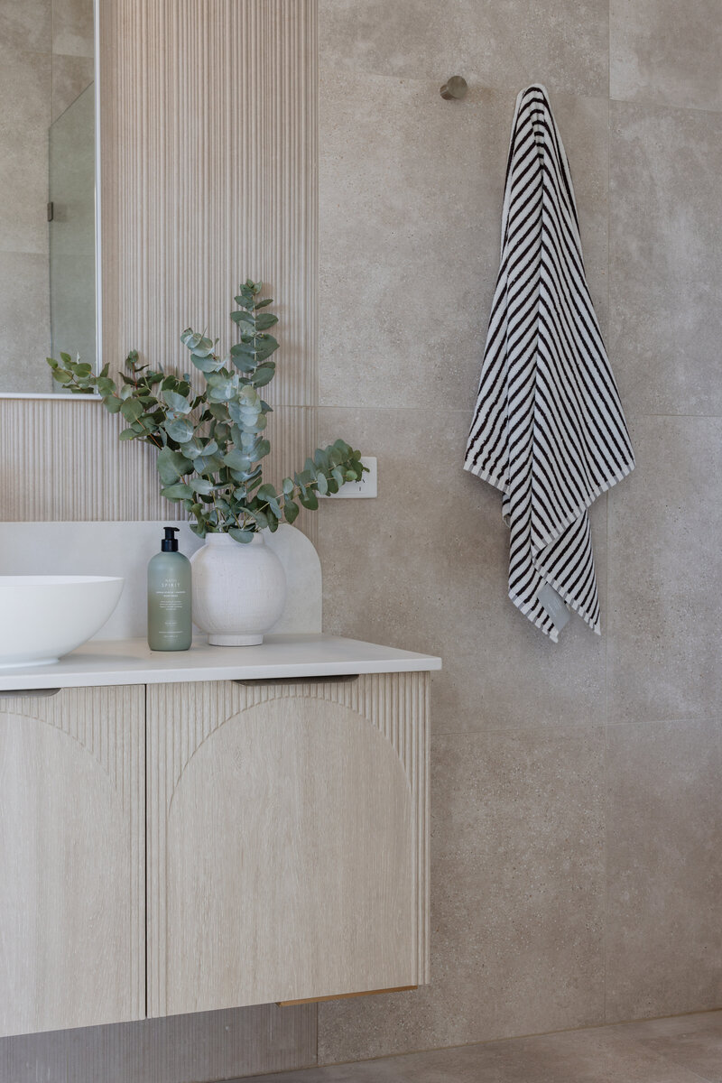 Minimalist bathroom featuring a light wood vanity, white sink, and striped towel. The space is elegantly accented with eucalyptus in a vase, showcasing the touch of bespoke interior design Newcastle NSW is celebrated for.