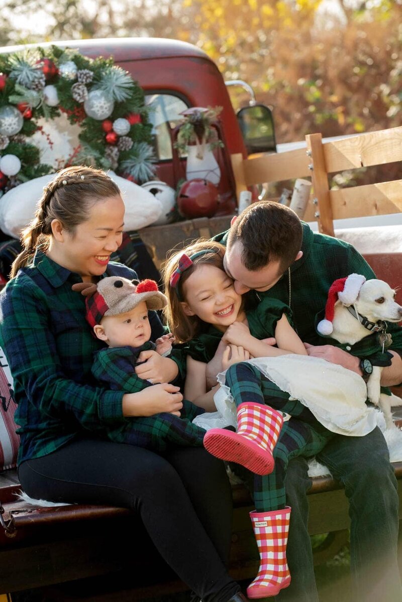 holiday family  in matching green plaid pajamas. Mom and dad laugh with daughter and young son and little dog while sitting on a vintage red ford truck.