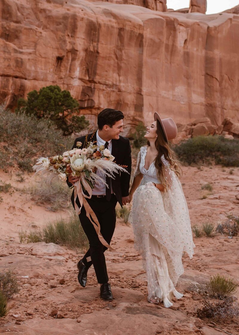 Bride and groom walking hand in hand through the red rock formations of Garden of the Gods in Colorado, with the bride wearing a boho dress and hat and holding a large floral bouquet during their intimate Colorado elopement