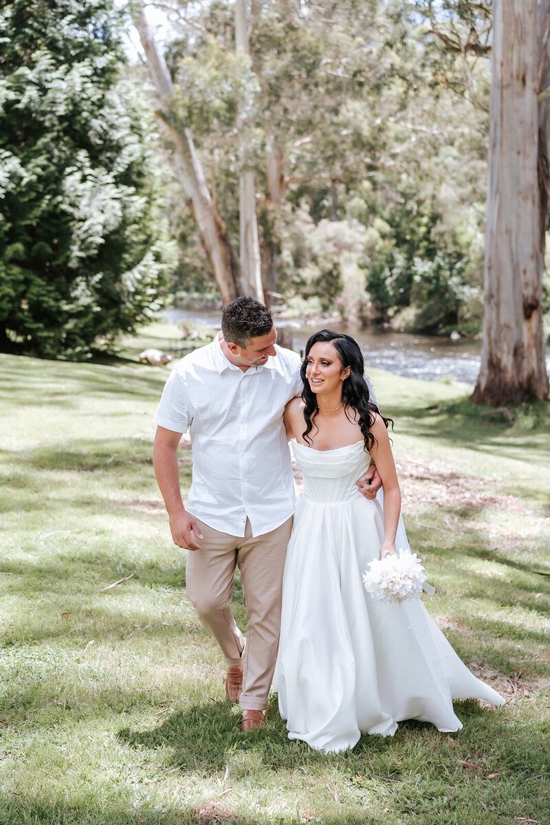 A bride in a white dress and a groom in a whie shirt and brown pants walking along the grass by the Yarra river.