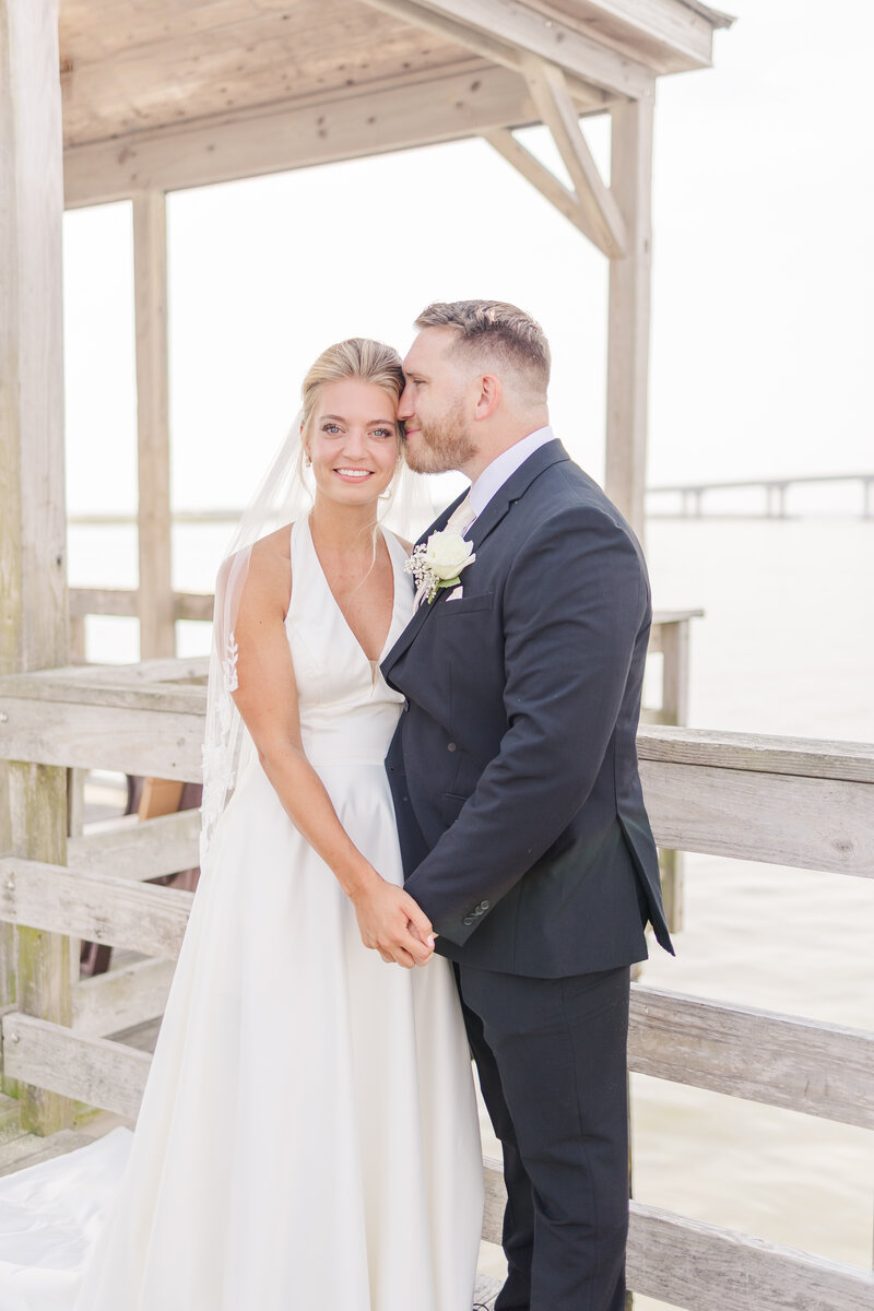 A groom in a black suit nuzzles his bride while standing on a wooden dock