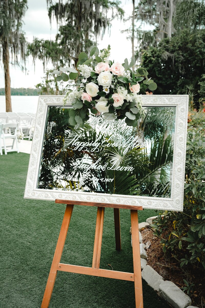 Mirror wedding sign with floral arrangement on a wooden easel.
