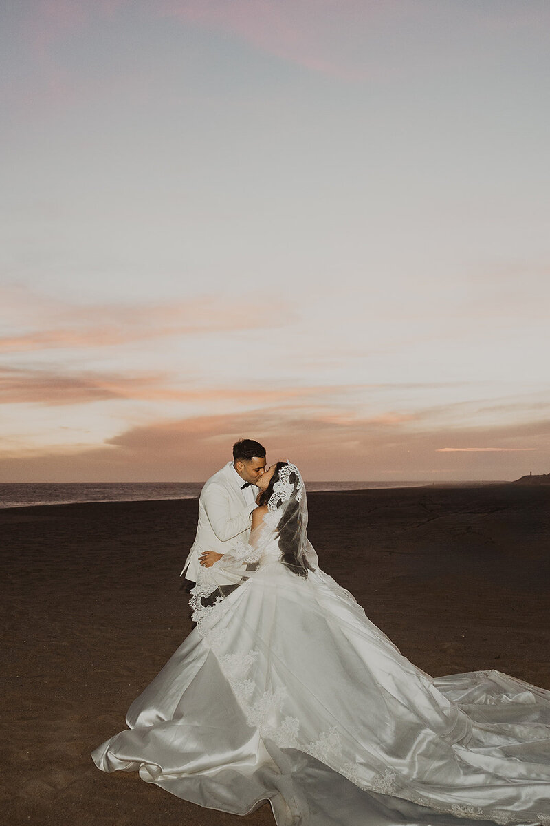 Bride and groom wearing wedding dress on beach kissing with sunset in background