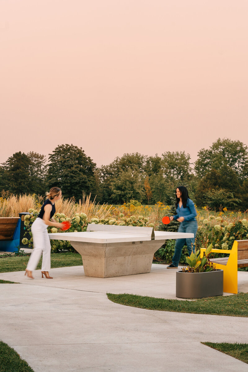 Two women playing ping pong  at an outdoor lounge surrounded by lush flowery bushes and trees
