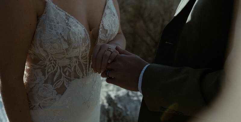 Close-up shot of a bride and groom holding hands during an emotional moment. The bride wears a fitted lace gown with floral embroidery and a deep V neckline, her engagement and wedding rings softly catching the light. The groom, dressed in a dark suit jacket, gently clasps her hands in his, their fingers interlaced.