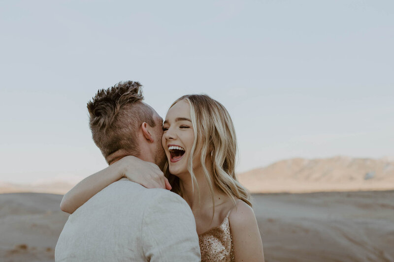Bride wearing gold dress and groom wearing tan suit laughing while hugging and laughing at the Sand Dunes during their elopement.