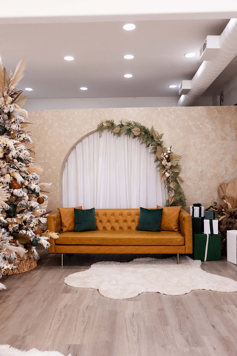 Boho holiday photography studio set with a wood arch wall furnished with a leather couch, Christmas Trees, and presents.