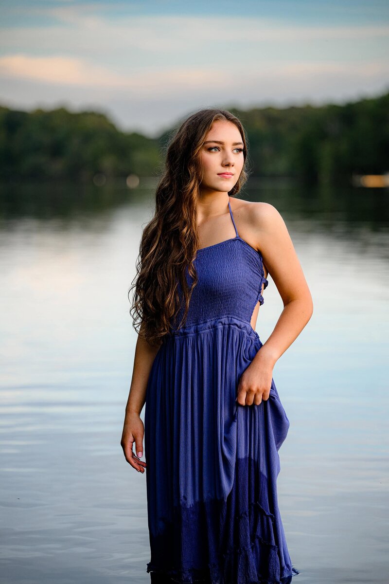senior stands in water during her photo session wearing a blue dress and looking off toward the sun