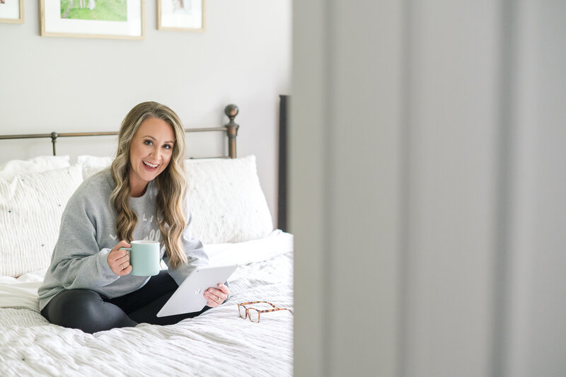 Jamie Fisher sitting on bed in sweatshirt holding mug and iPad smiling at camera