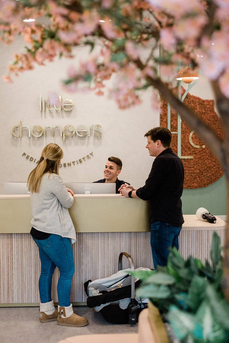 A mother and father speak with the male receptionist at Little Chompers Pediatric Dentistry on Clark Street. The faux floral blooms of the tree in the patient waiting area frame the photo in the foreground.