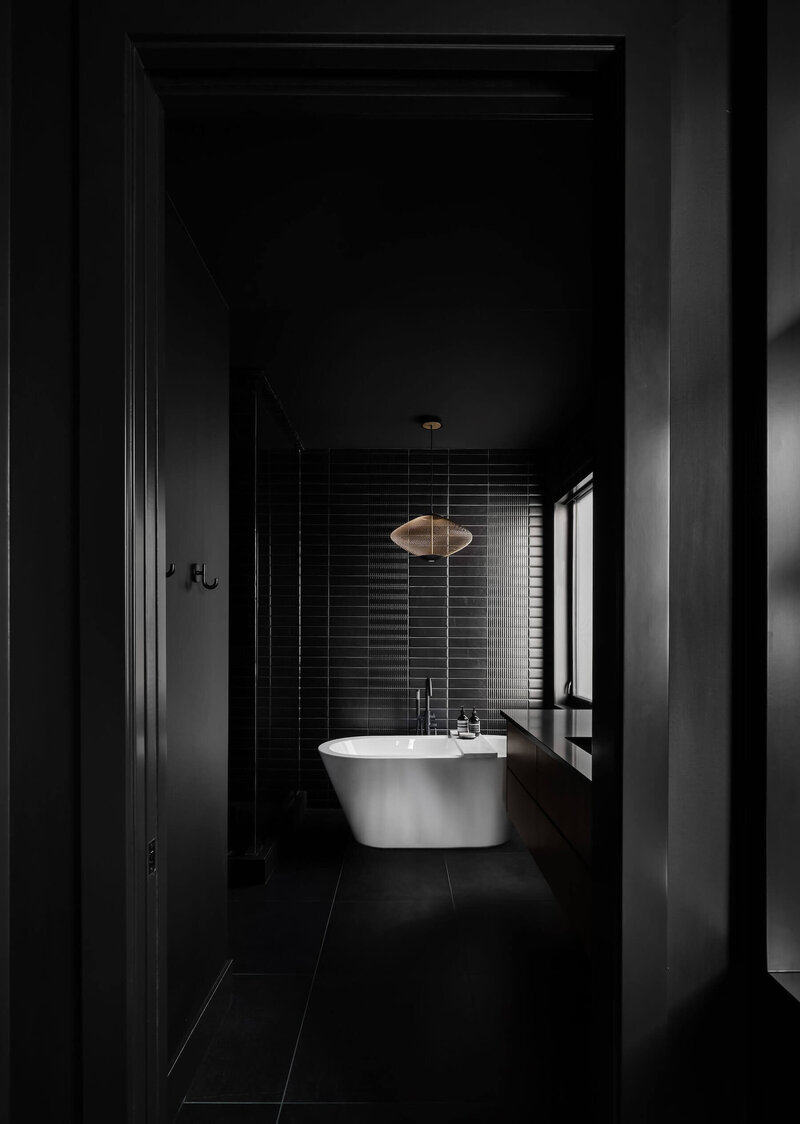 A dramatic black bathroom with black subway tile, gold light feature and free standing tub. 