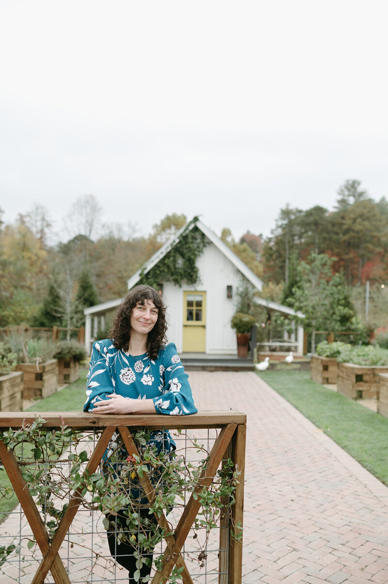 A portrait of artist and designer Skye McNeill. Skye wears a dark teal floral print dress and stands at the entrance gate to a farm. A cute yellow dutch door and duck house are behind her. Photograph by Callie Lynch.