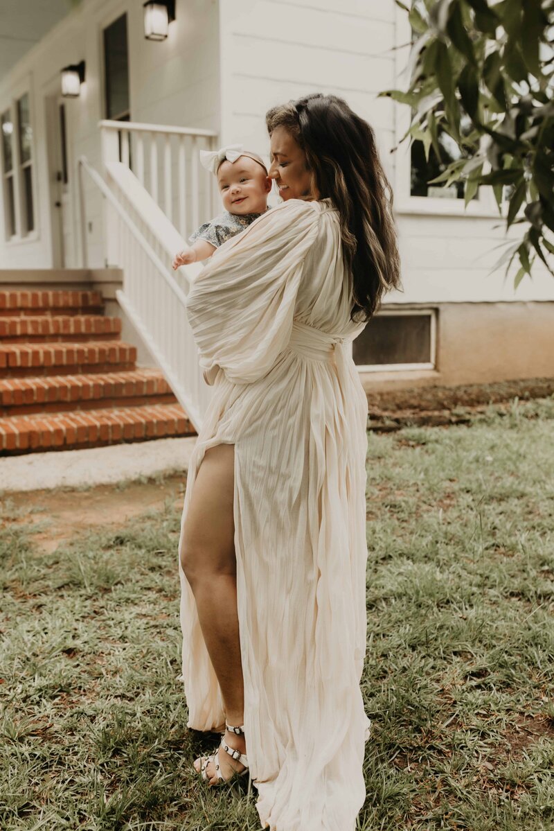 A woman in a flowing cream dress stands on grass, holding a smiling baby in her arms, in front of a white house with red brick steps—captured by Macon family photographer, Fire Family Photography.