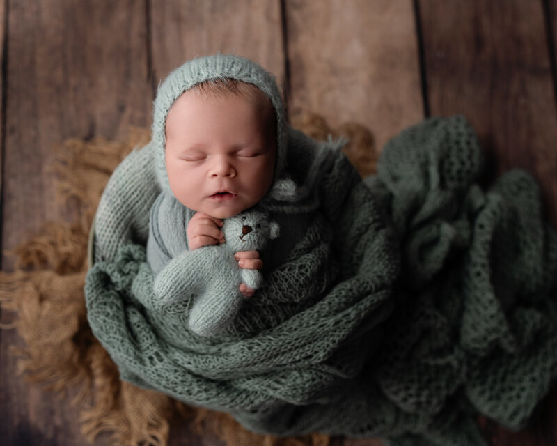 A cozy newborn portrait featuring soft sage knits and a beloved teddy companion—styled with rustic charm.