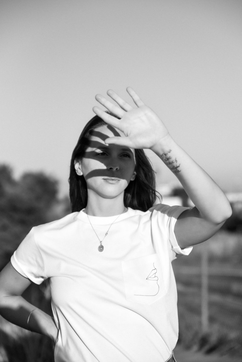 Black and white self portrait of a brunette woman in a Hies  t-shirt holding up her hand to block the sun
