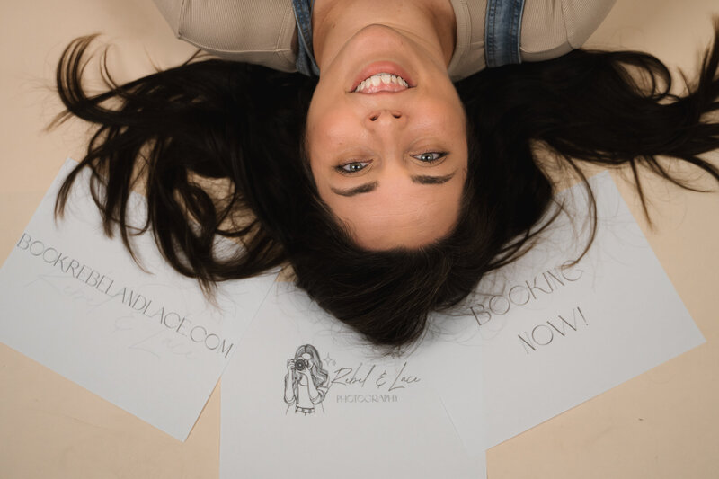 Image of a brunette looking into the camera, laying on her back surrounded by papers advertising Rebel and Lace.