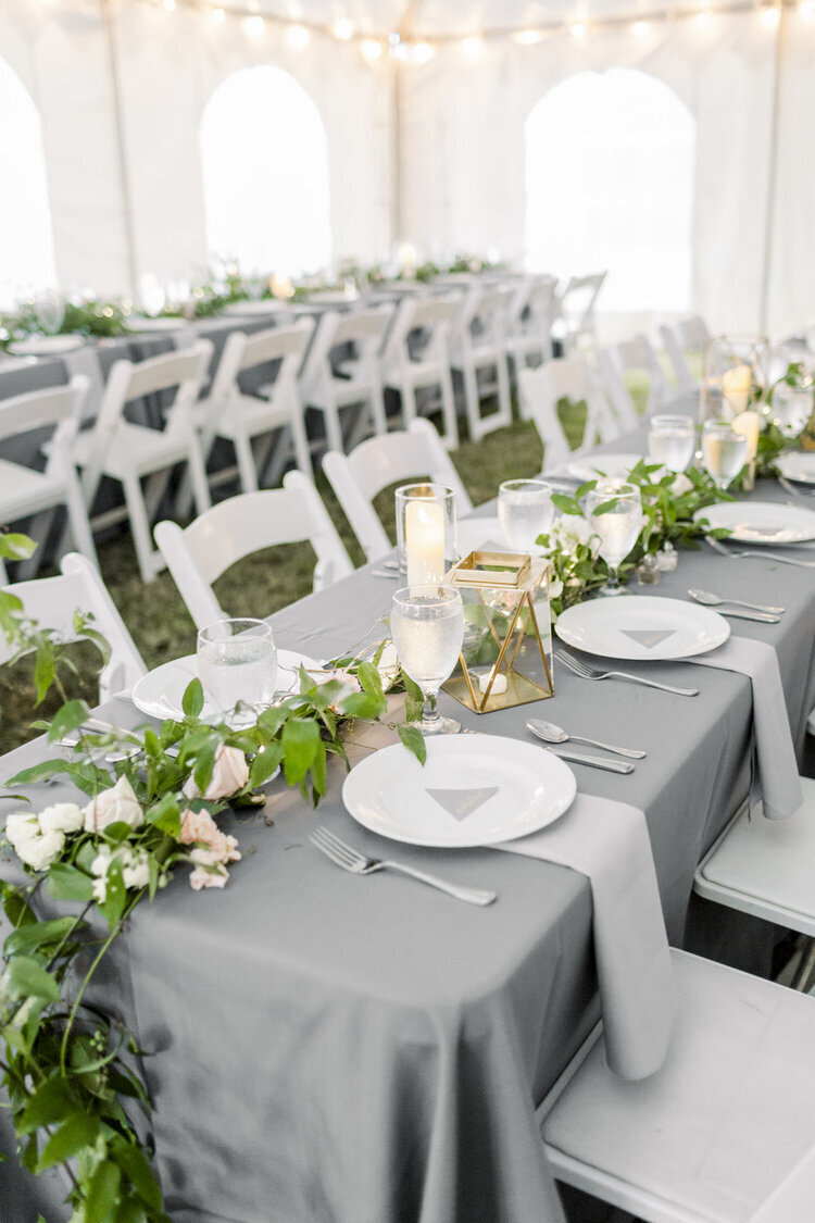 outdoor wedding reception with long kings tables with grrey linens and greenery with gold accents and candles and white plates with silverware and white chairs at the glasgow farm in fredericksburg, virginia planned by cait and co events