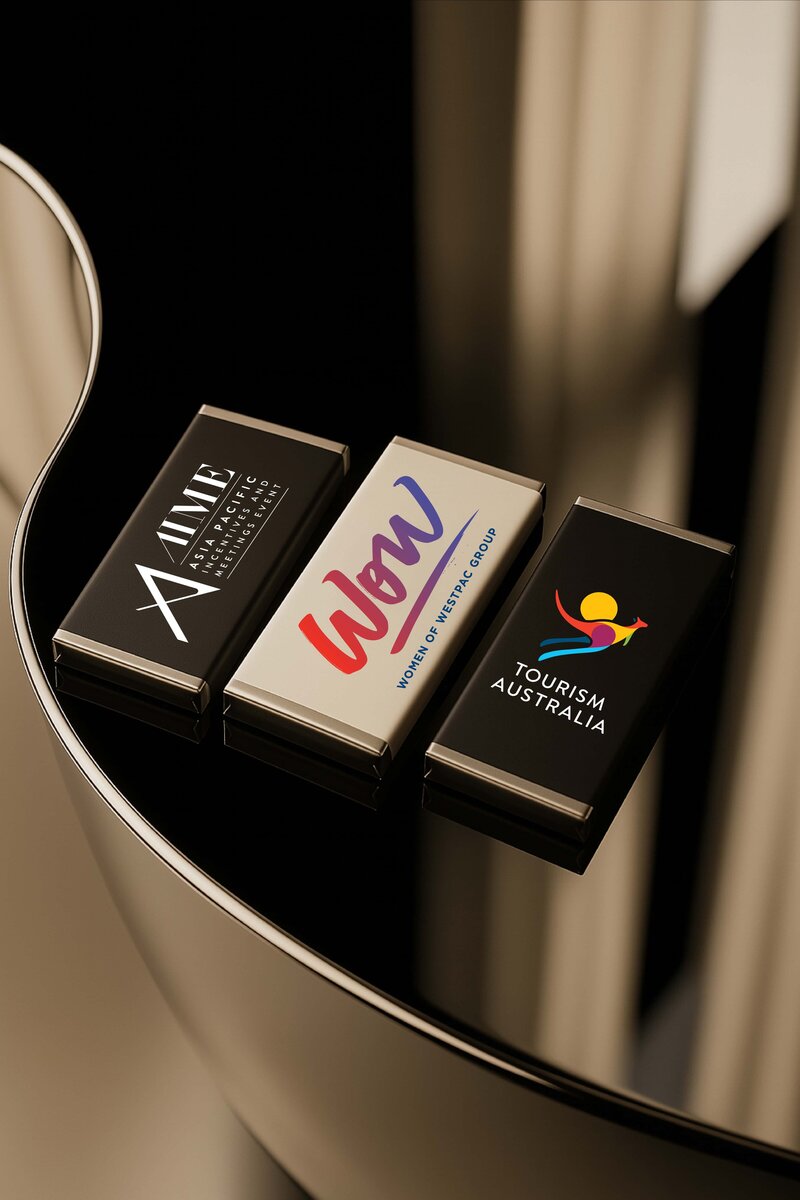 Personalised corporate chocolate bars featuring AIME, Women of Westpac, and Tourism Australia logos, designed by Barilliant with luxe silver foil ends, styled on a reflective black surface.