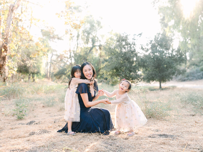 A mom in a navy dress kneels in a sunlit forest, smiling warmly. Two young girls in matching pastel pink dresses hug and hold her hands. The scene is joyful and serene.
