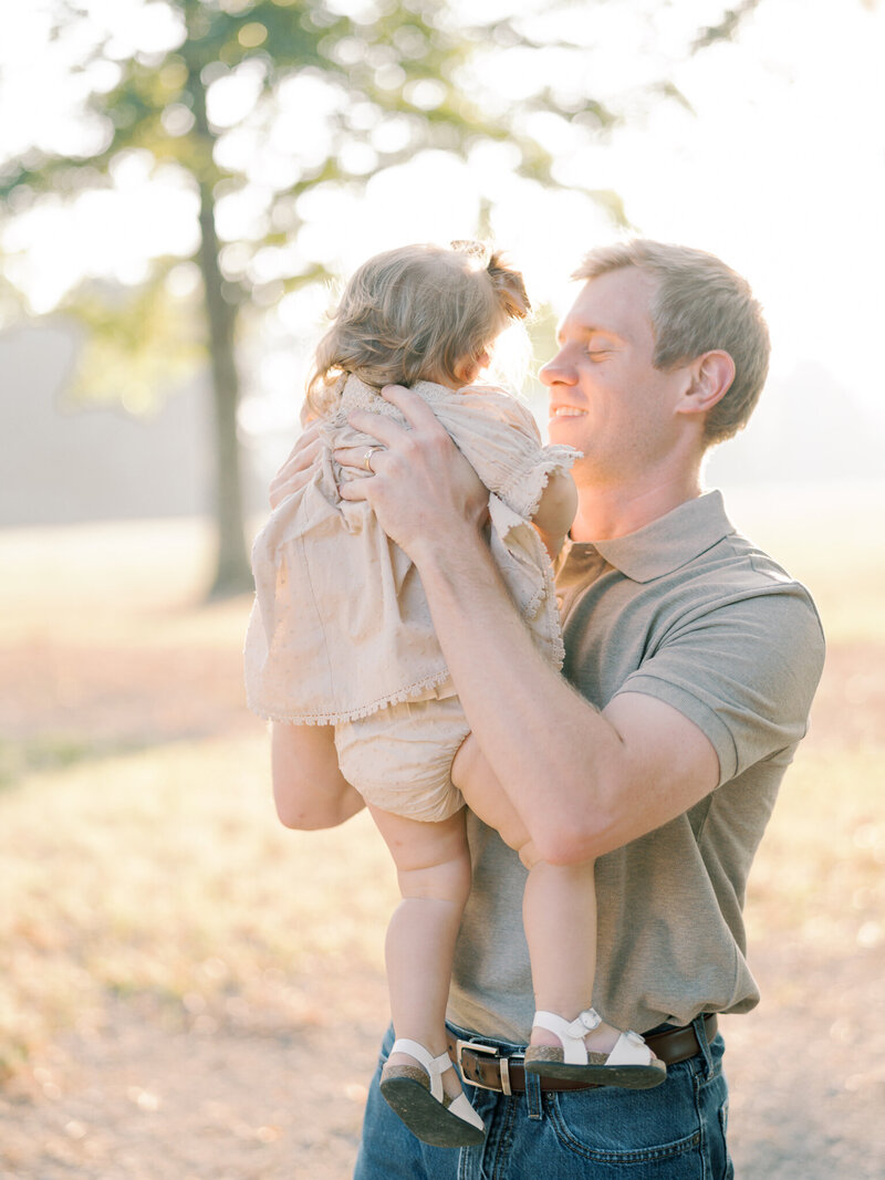 Blonde dad in brown shirt and jeans lifts his young, blonde daughter into air, smiling at her, taken by Little Rock photographer Bailey Feeler