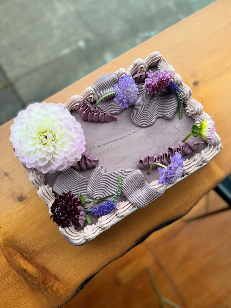 Rectangular lavender-frosted gluten-free cake decorated with fresh flowers and piped buttercream details, handcrafted at Grain Artisan Bakery in Snohomish, WA.