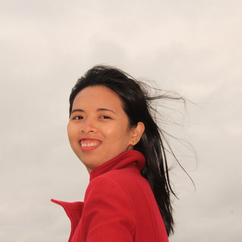 Smiling woman in a bright red coat with wind in her hair, standing outdoors against a cloudy sky backdrop