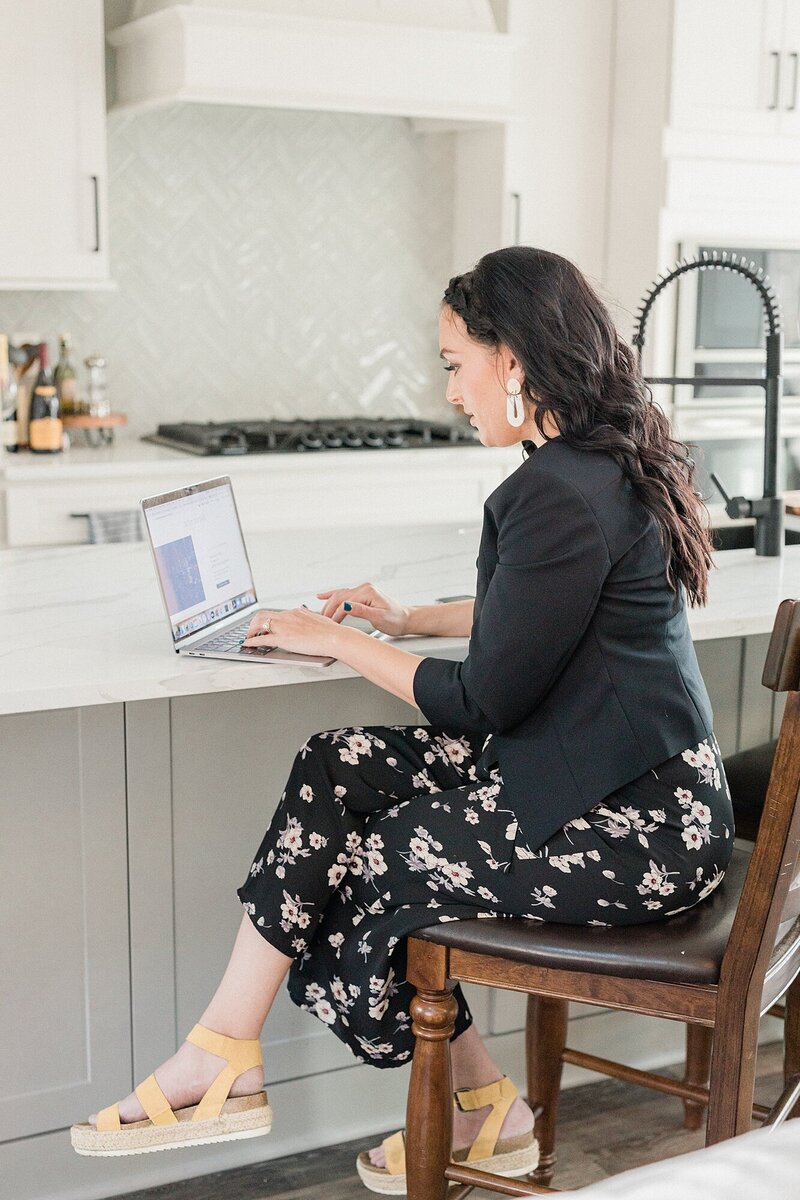 woman with black hair sitting at a kitchen island working on a macbook pro and smiling