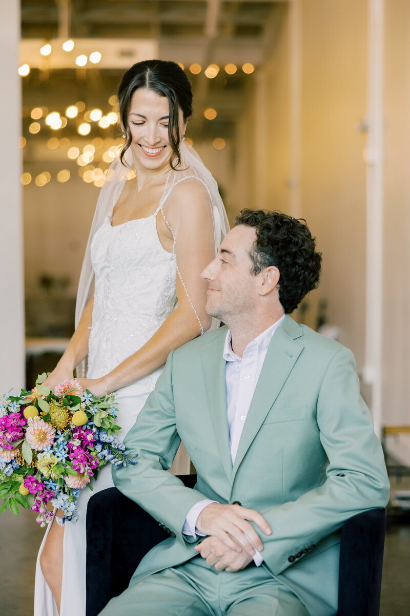 Editorial wedding portrait indoors; bride in beaded gown and veil stands behind partner in mint suit, colorful bouquet.