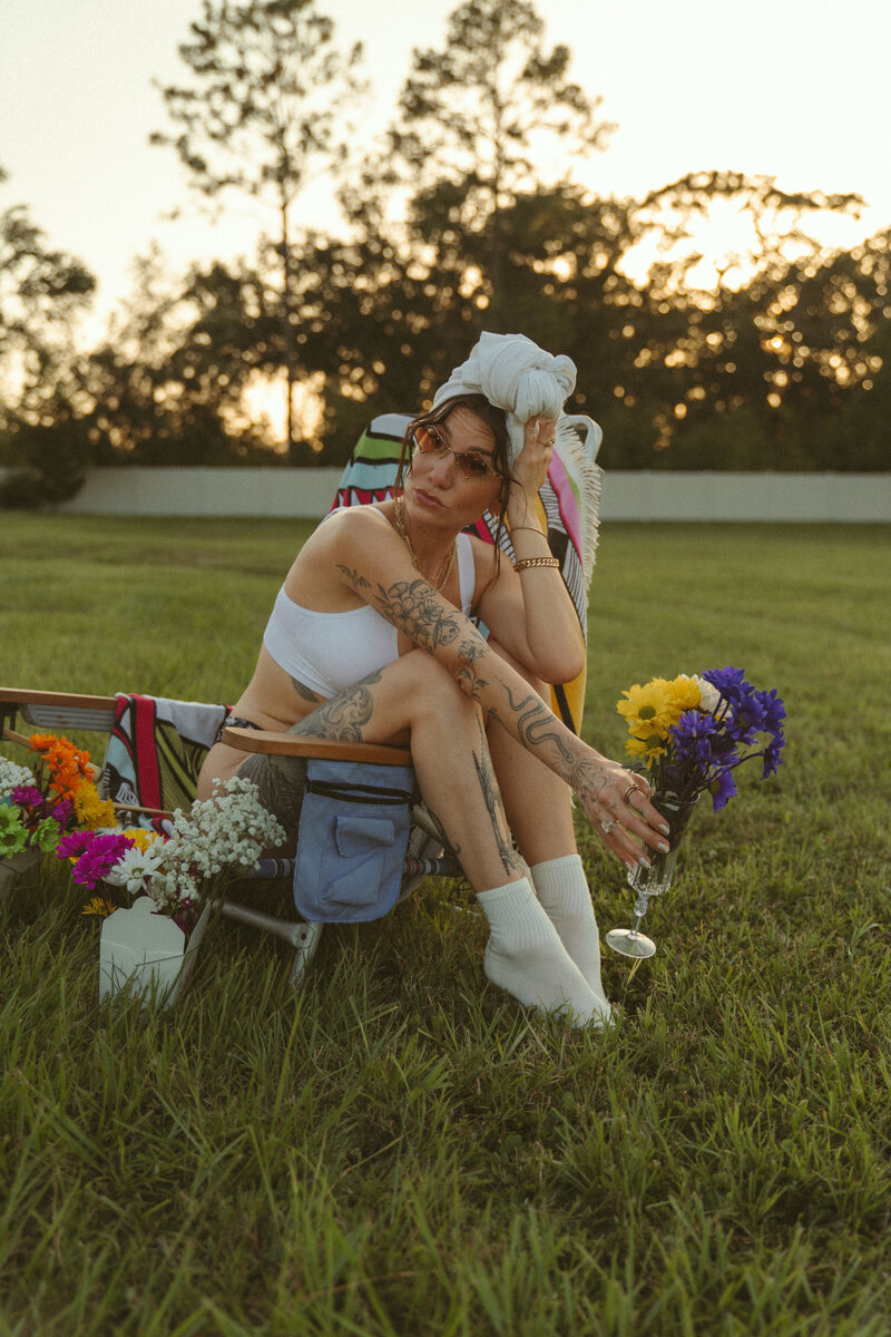 Tattooed woman sitting on a colorful lawn chair in a grassy field at sunset, holding a wine glass filled with vibrant flowers, wearing sunglasses, white crop top, and a head wrap, styled in a bold editorial aesthetic.