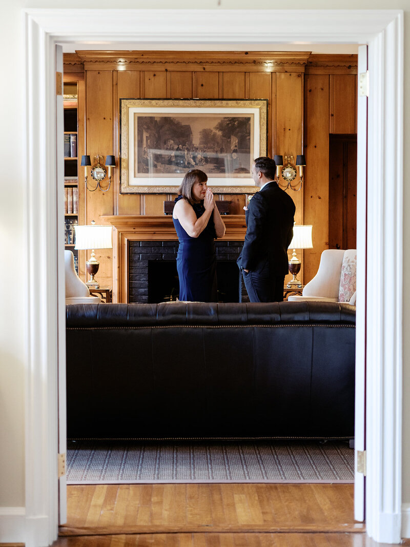 Cameron shares an emotional first look with his mother at Boxwood Estate. He stands in a beautifully landscaped area, looking at his mother as she smiles, holding back tears. Cameron is dressed in a sharp suit, while his mother wears an elegant dress. The soft natural light and the estate's picturesque setting create a heartfelt and intimate moment between mother and son, capturing the bond they share before the wedding.