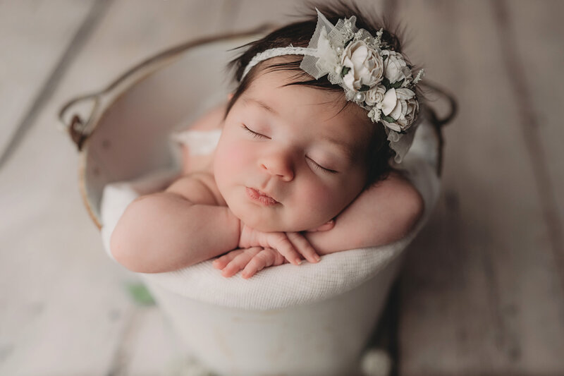 sleeping newborn baby girl wearing white flower bow in  hands on chin pose inside of bucket over rustic white wood plank backdrop