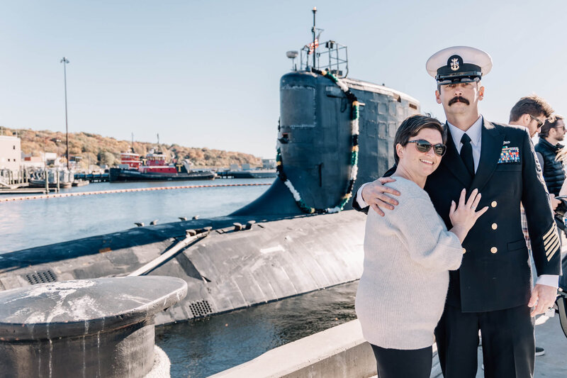 Man in Navy Chief uniform and woman looking at camera in front of recently returned submarine USS North Dakota at SUBASE New London in Groton, CT.