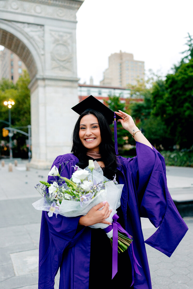 NYU graduate smiling in front of the arch at Washington Square Park while wearing cap and gown and holding a bouquet of flowers.