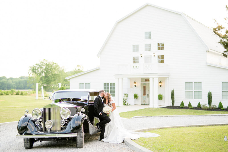 Couple kisses by vintage car and white barn.
