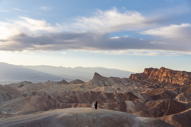 A wide landscape shot of a queer couple standing among golden rocky hills as the sun sets behind mountains