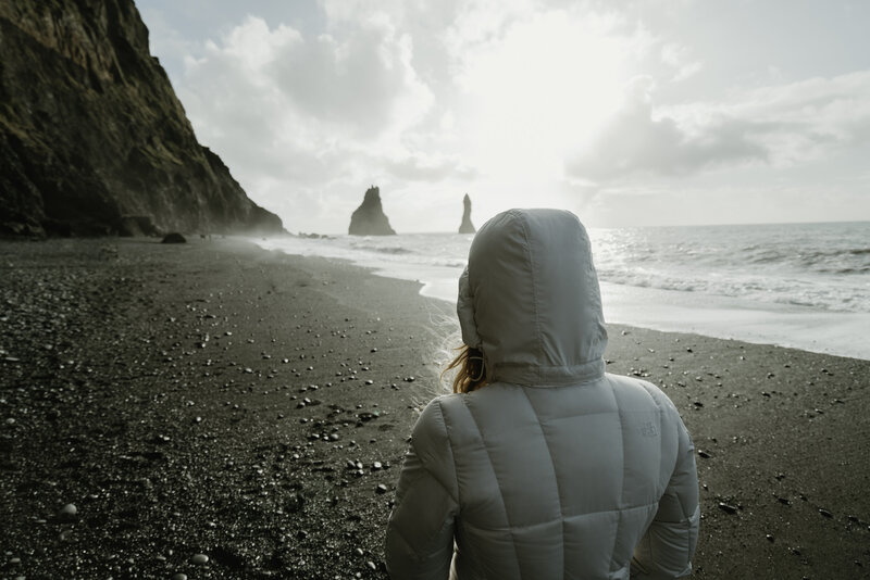 A woman in a hooded white coat stands facing the ocean on a pebbled beach under a bright sky. In the distance, jagged sea stacks rise from the sea, creating a dramatic landscape. The sun, partially obscured by clouds, casts a soft light across the scene, highlighting the textures of the dark cliffs and the foamy sea waves.
