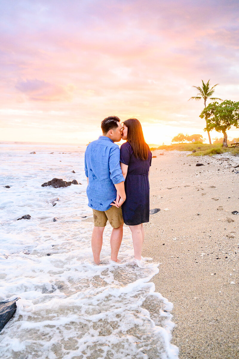 couple wearing navy and blue face eachother and kiss while the waves crash under their feet in the sand