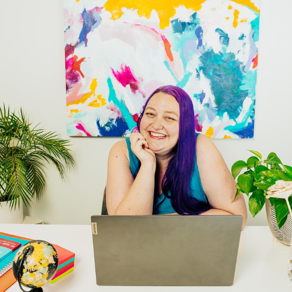 A woman with vibrant purple hair sits at a desk, smiling at the camera. She is wearing a blue sleeveless top, with her hand resting on her chin, and the background features a colorful abstract painting along with some greenery and books on the desk.