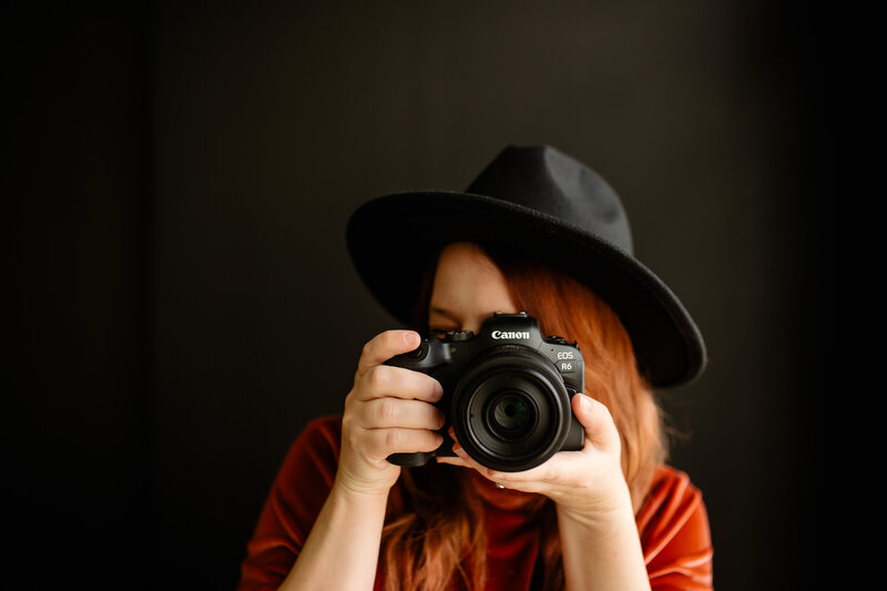Photograph of a woman with the camera covering her face, wearing a black hat and an orange T-shirt