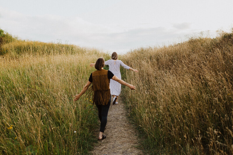 Kaksi aikuista siskosta kävelee heinäniityn poikki polkua pitkin ja levittävät käsiään koskettaen heiniä Suomenlinnassa Helsingissä