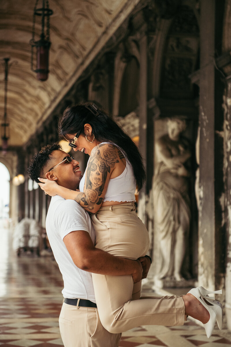 This stunning photo showcases a newly engaged couple in San Marco Square in Venice, with the man joyfully lifting his partner up in the air as they both beam with happiness. The historic porch overhead and the vibrant energy of the bustling square add to the enchanting atmosphere of this picture-perfect moment. The image captures the pure joy and love shared between the couple in a timeless scene that will be cherished forever