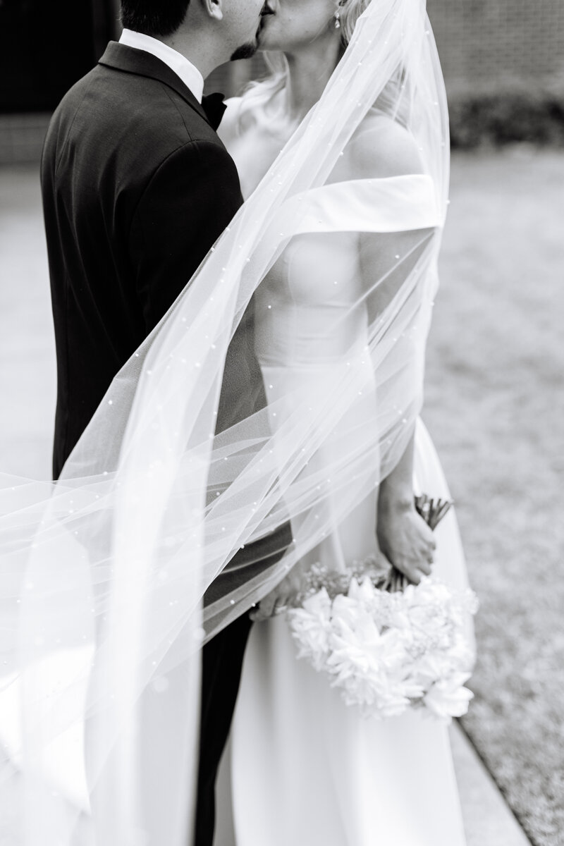 Black and white photo of bride and groom kissing with bride's veil blowing in the wind