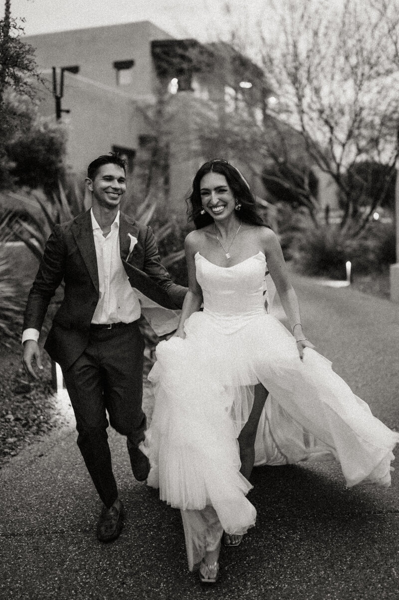 A couple celebrates their elopement in Las Vegas, with neon casino signs lighting up the background. One partner, wearing a short white dress and veil, smiles brightly while being lifted by the other, showing off their ring.