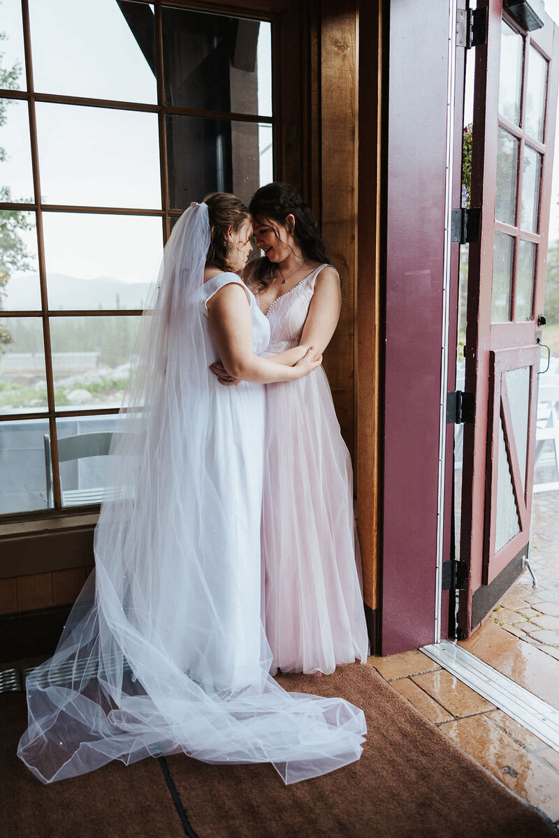 Two brides standing close together in a corner on their wedding day.