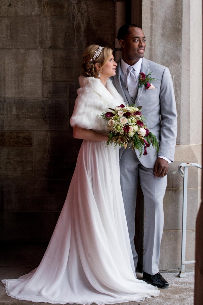 a bi-racial couple stands next to a window for their wedding photography. The bride wears a fur shawl, holding maroon and white flowers