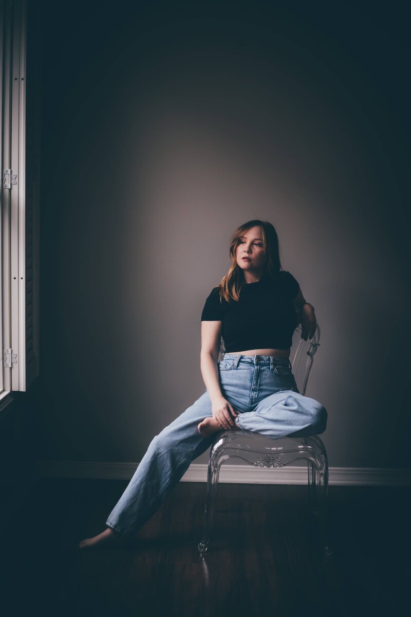 A woman with long hair sits on a transparent chair in a dimly lit room. She wears a black top and blue jeans, with one leg crossed on the chair and the other on the floor, gazing off to the side, contemplating whether to hire an online business manager for her growing startup.