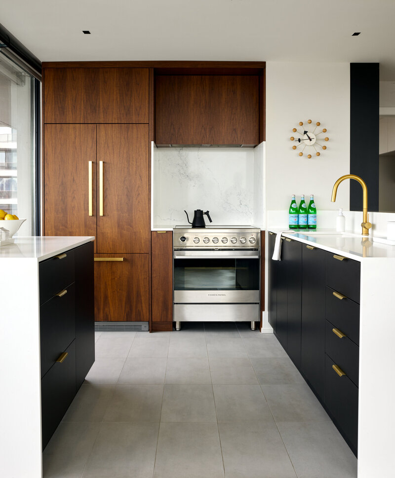 Before and after transformation of a plain white kitchen into a bright kitchen featuring luxury black and walnut cabinetry, herringbone white-washed oak floors, a stainless steel stove and oven, decorative wall clock, and gold accents.